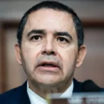 A middle-aged man wearing a dark suit speaks into a microphone at a formal hearing, with a wooden panel background behind him.