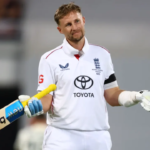 An England cricketer in a white Test uniform stands on the field holding his bat in one hand and his helmet in the other, giving a small shrug with a calm, almost playful expression after completing a milestone innings.
