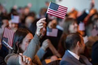 A crowd of people sit together at a ceremony, many of them holding small American flags, with one person in the foreground raising a flag in the air.