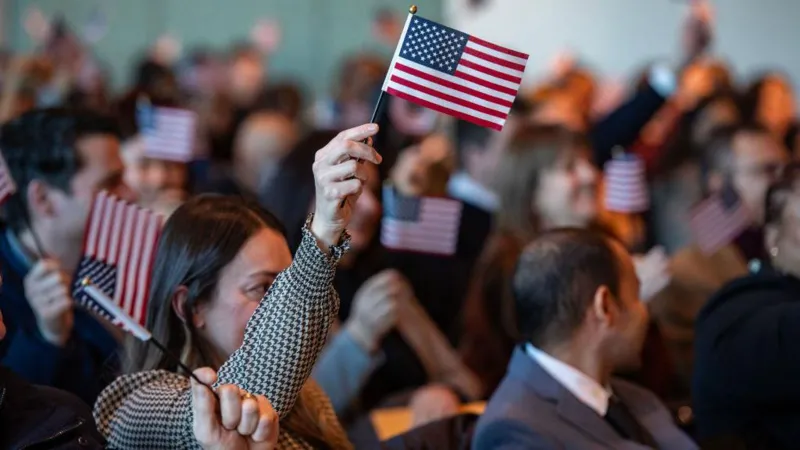 A crowd of people sit together at a ceremony, many of them holding small American flags, with one person in the foreground raising a flag in the air.