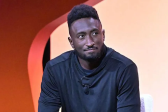 A man in a black long-sleeve shirt sits on stage against an orange backdrop, looking thoughtful during a tech-related discussion.