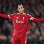 A soccer player wearing a red Liverpool FC kit, identified as Virgil van Dijk, stands on the pitch with his arms stretched out to his sides in a gesture of confusion or exasperation. He is wearing the captain's armband and his jersey has the standard chartered sponsor logo. The background is a slightly blurred, dark stadium crowd.