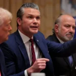 A group of men in formal suits sit at a conference table. The man in the middle is speaking passionately and pointing forward, while the man to his left listens closely. Two other men sit on the right, watching the discussion.